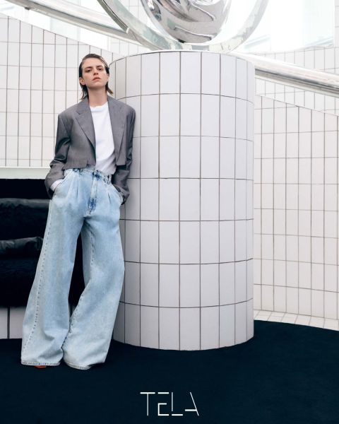Persona appoggiata a una colonna piastrellata di bianco che indossa un blazer grigio, una t-shirt bianca e jeans azzurri oversize. Il pavimento è nero e l'ambiente è minimalista con decorazioni moderne. In basso c'è la parola "TELA".
