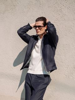 Un uomo che indossa occhiali da sole, una giacca blu navy e pantaloni abbinati è in piedi con le mani sulla testa davanti a un muro chiaro e strutturato.