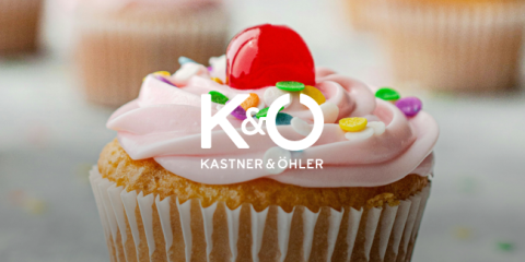 Im Fokus steht ein Cupcake mit rosa Zuckerguss, einer Kirsche obendrauf und bunten Streuseln, im Hintergrund sind mehrere verschwommene Cupcakes zu sehen. Über dem Bild sind in Weiß die Buchstaben „K & O“ und der Text „KASTNER & ÖHLER“ zu sehen.