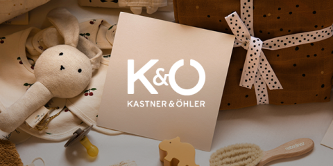 Eine Sammlung von Babyartikeln, darunter ein Plüschhase und eine Holzbürste, umgibt eine Geschenkkarte von Kastner & Öhler. Die Karte zeigt das K&O-Logo in Weiß auf beigem Hintergrund.