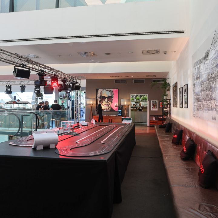 Stanza con un grande tavolo da corsa per slot car al centro. Sedute imbottite fiancheggiano una parete e un murale con un paesaggio urbano decora un'altra. Le luci a soffitto creano un'atmosfera accogliente e un poster incorniciato è appeso alla parete di fondo.