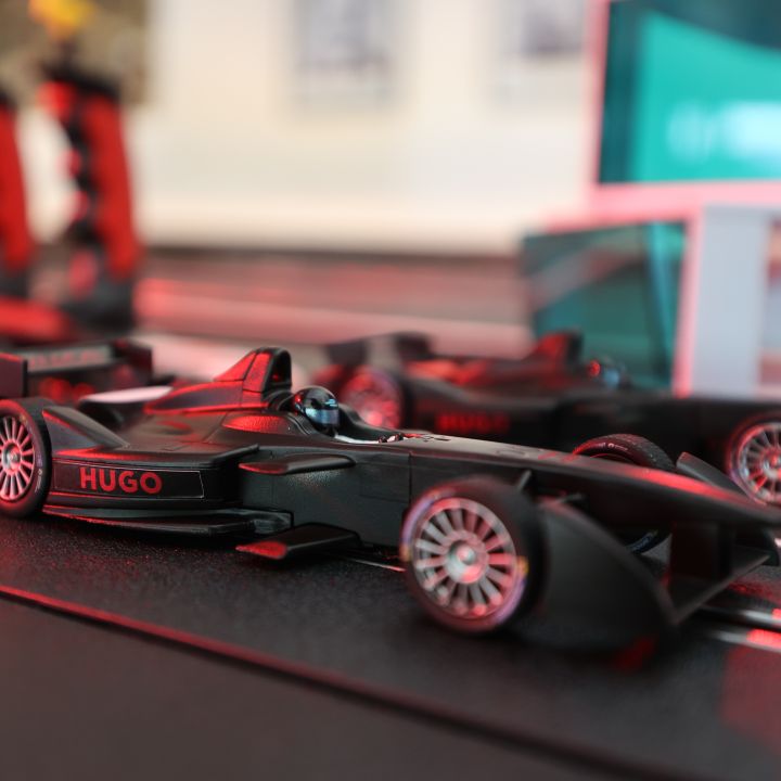 Primo piano di un'auto di Formula 1 in miniatura nera su un circuito con il nome "Hugo" sul lato. L'auto ha un design elegante con pneumatici visibili e sullo sfondo si intravede un'altra auto leggermente sfocata.