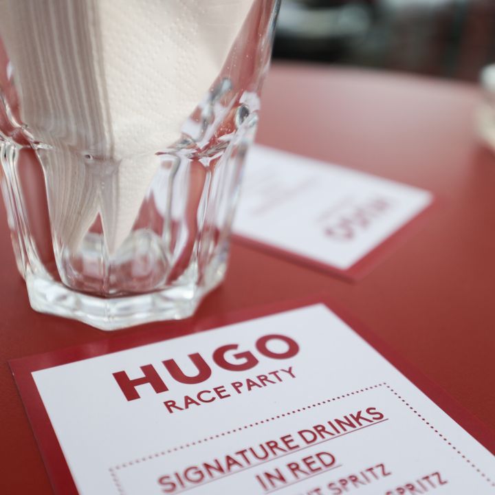 Primo piano di un tavolo apparecchiato in un ristorante con un bicchiere contenente tovaglioli piegati. Sul tavolo c'è un menu con l'intestazione "Hugo Race Party" e un elenco di "Signature Drinks" tra cui "Vermut Spritz" e "Crodino Rosso Spritz".