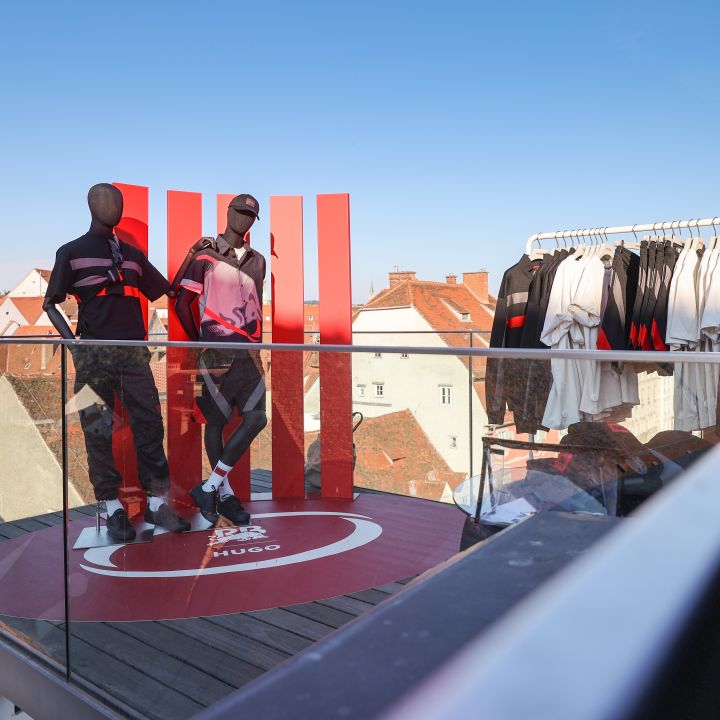 Due manichini in abbigliamento sportivo si trovano su un'esposizione sul tetto con ringhiera in vetro. A destra c'è un espositore con abbigliamento simile. Sullo sfondo si vedono tetti e un cielo azzurro limpido.