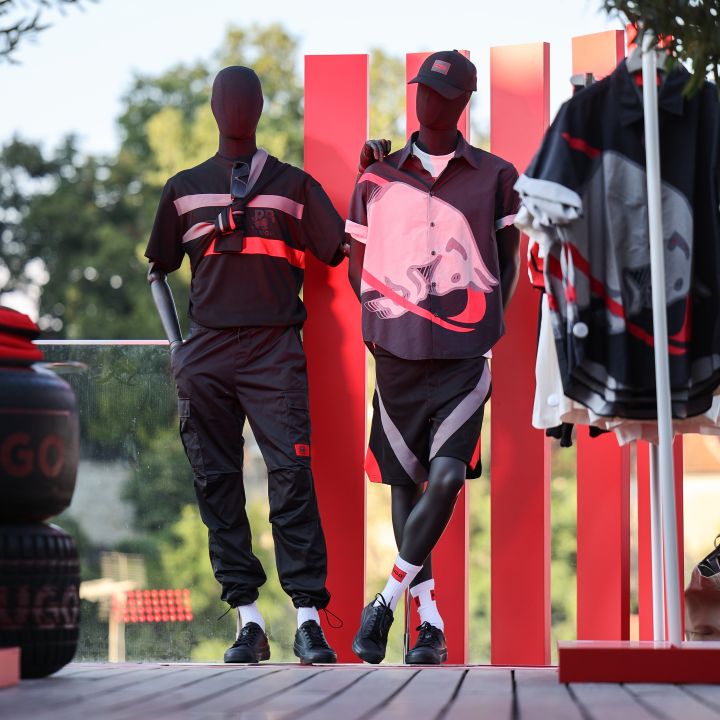 Due manichini in abiti sportivi si trovano su un ponte di legno davanti a uno sfondo a strisce rosse e bianche. Il manichino a sinistra indossa abbigliamento casual nero e rosso, mentre il manichino a destra indossa un completo sportivo nero e rosso con un cappello. Sullo sfondo si vedono degli alberi.