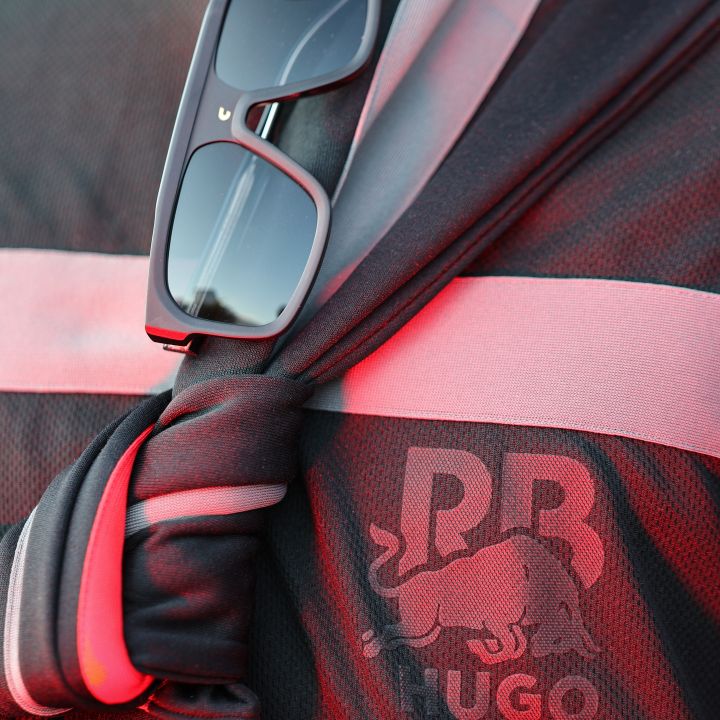 Primo piano di una maglia sportiva nera e rossa con i loghi "RB" e "Hugo" con un toro. Un paio di occhiali da sole neri sono infilati nel colletto della maglia, che presenta un dettaglio in tessuto annodato. L'illuminazione proietta una calda tonalità rossa sull'indumento.