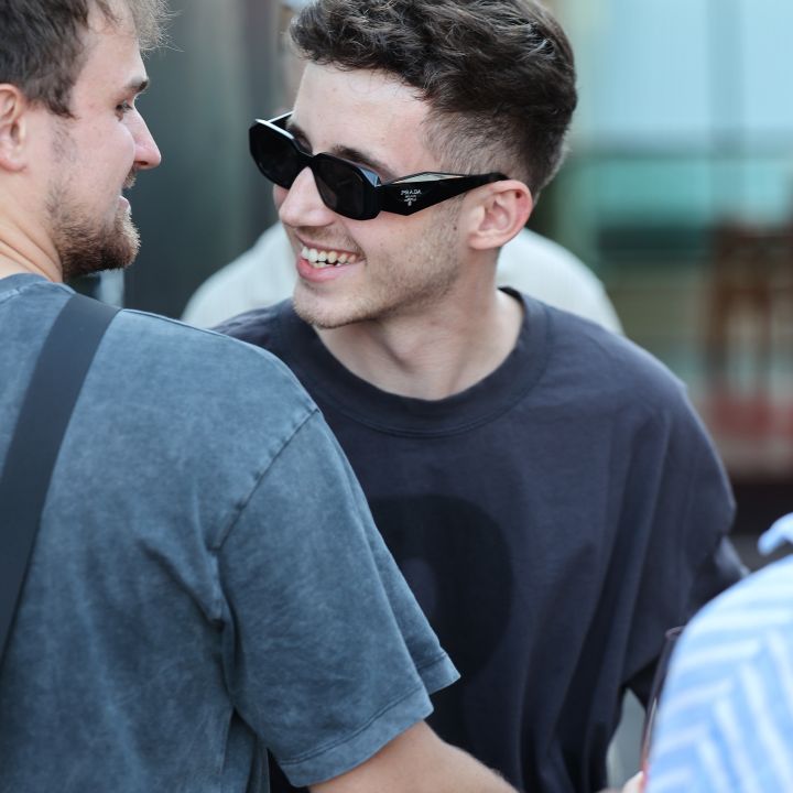 Due giovani uomini si sorridono all'aperto. Uno indossa occhiali da sole neri e una maglietta scura, mentre l'altro ha la barba chiara e una camicia grigia. Sembrano salutarsi o trascorrere un momento amichevole insieme.