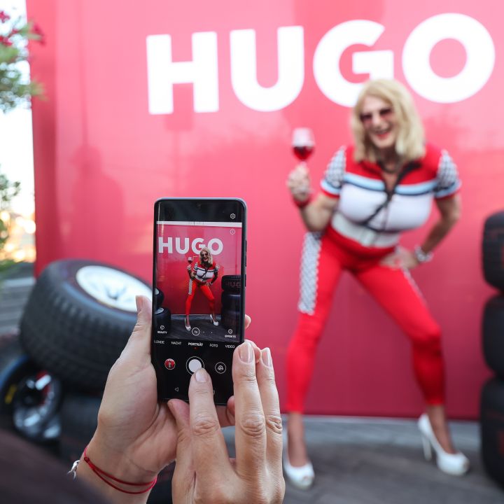 Persona in abito rosso e bianco posa con un bicchiere di vino davanti a uno sfondo rosso con il testo "HUGO". Un'altra persona scatta una foto con un telefono. Sul lato destro sono impilati dei pneumatici.