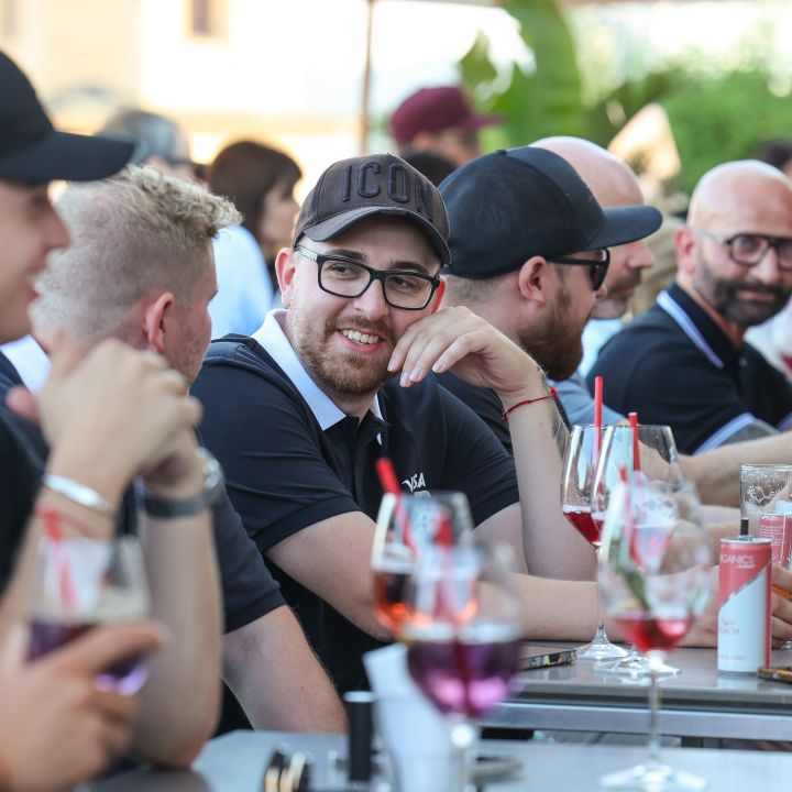 Un gruppo di persone è seduto a un lungo tavolo all'aperto e gusta bevande in bicchieri con liquido rosso. Sono vestiti in modo informale, alcuni indossano cappelli e occhiali da sole e sembrano immersi in una conversazione e sorridono.
