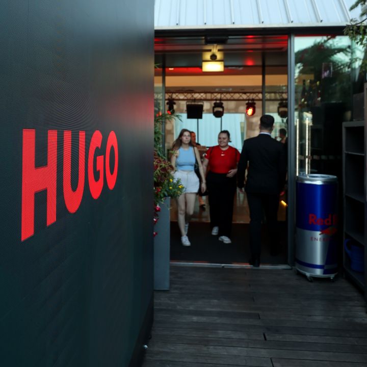 Ingresso a un locale con un grande cartello "HUGO" su una parete. Tre persone entrano: una donna in abito bianco, un uomo in camicia rossa e un uomo in abito scuro. A destra c'è un frigorifero Red Bull e sopra un tetto spiovente.