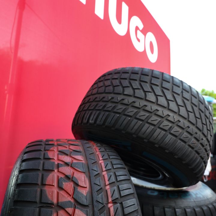 Tre pneumatici impilati con evidenti tracce di battistrada si trovano di fronte a un muro rosso su cui è scritta la parola "HUGO" in grandi lettere bianche.