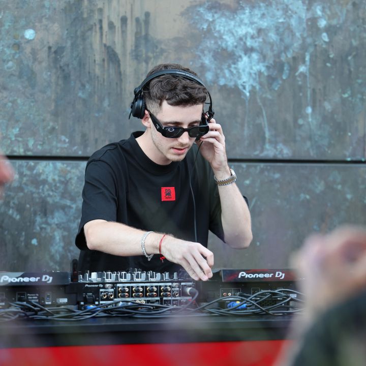 Un DJ con occhiali da sole e cuffie è concentrato a regolare i controlli di un mixer Pioneer DJ. Sta suonando musica all'aperto, con persone sfocate in primo piano e un muro strutturato sullo sfondo.