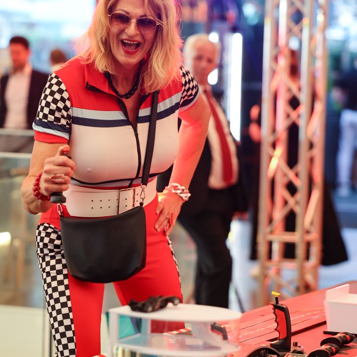 Donna in abito a quadri rosso e nero sorride entusiasta indicando una pista da corsa in miniatura. Indossa occhiali da sole e un marsupio nero. Sullo sfondo si intravedono luci brillanti e altre persone in un locale.