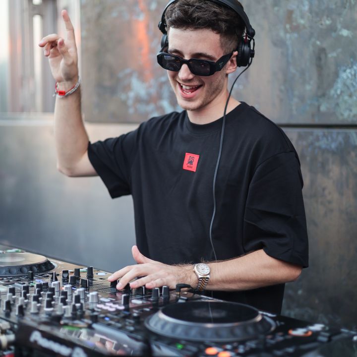 Un DJ con occhiali da sole e cuffie suona musica con energia a un mixer. Alza una mano e sorride. Indossa una maglietta nera con un motivo rosso. Sullo sfondo si vede una parete metallica strutturata.