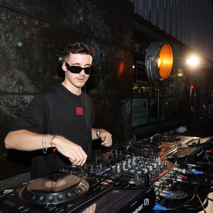 Un DJ con occhiali da sole e camicia nera si esibisce a un evento all'aperto. Utilizza una console con vari controlli e pulsanti. Sullo sfondo si vedono una parete strutturata e una luce rotonda il cui raggio illumina l'area.