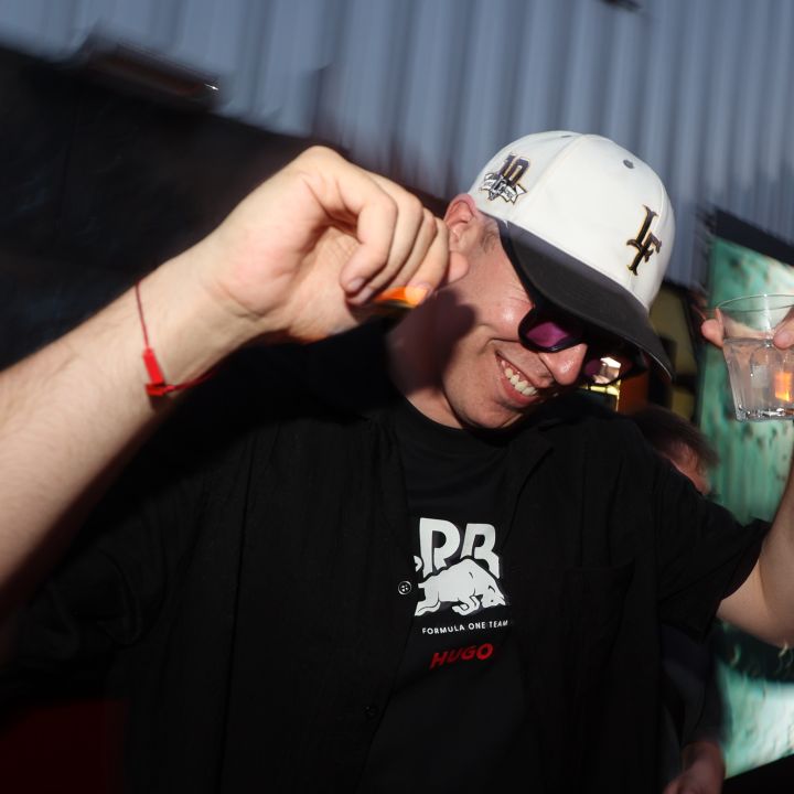 Una persona con camicia nera, occhiali da sole e cappello bianco sorride e balla, tenendo un drink in una mano. Lo sfondo è un murale sfocato su una struttura metallica che suggerisce un ambiente esterno.