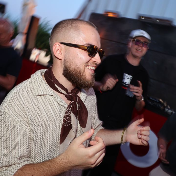 Un uomo con barba e occhiali da sole, che indossa una camicia a motivi e una sciarpa, balla allegramente a un evento all'aperto. Diverse altre persone sono in piedi intorno a lui e si godono anche la musica e l'atmosfera. Lo sfondo è leggermente sfocato.