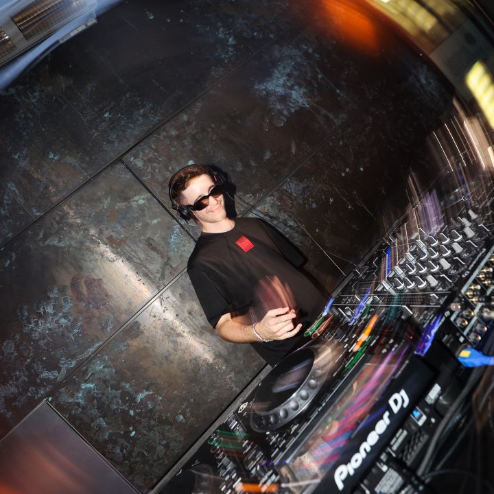 Un DJ con occhiali da sole e cuffie mixa a un mixer. La scena è illuminata da una luce calda e ci sono effetti di movimento sfocati che conferiscono energia dinamica all'immagine.