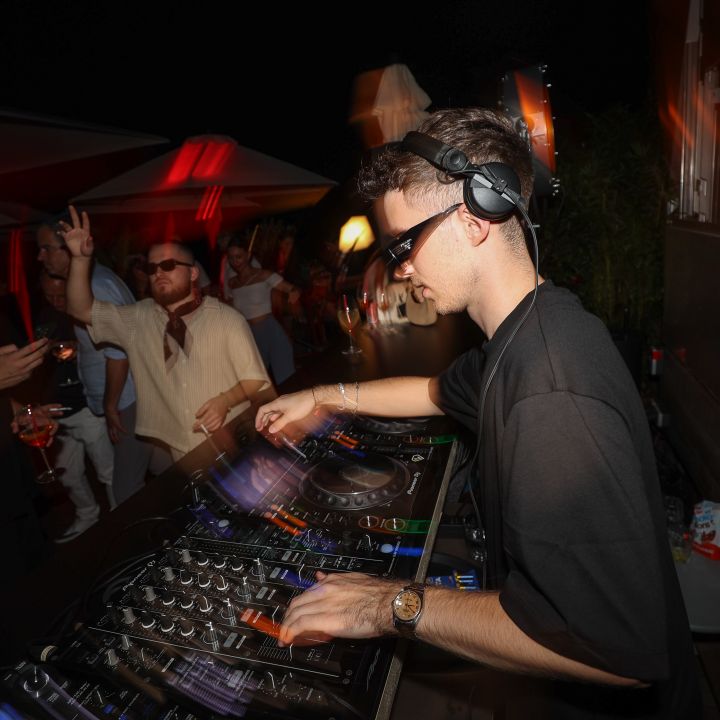 Un DJ con occhiali da sole e cuffie mixa musica su un mixer durante un vivace evento all'aperto. Due persone sullo sfondo si godono l'atmosfera, una tiene in mano un drink. Luci colorate e un cielo notturno scuro creano una vivace scena di festa.