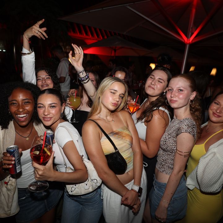 Un gruppo di amici posa per una foto durante un evento notturno. Sono vestiti in modo informale, alcuni tengono in mano delle bevande e sembrano divertirsi. Sullo sfondo si vedono luci rosse e grandi ombrelloni.