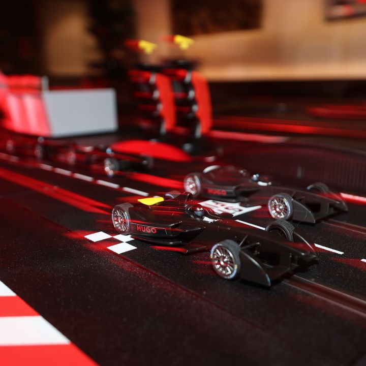 Primo piano di due auto da corsa giocattolo su una pista nera con segnaletica rossa e bianca. La scena include barriere e strutture rosse e nere che creano un ambiente di corsa in miniatura.