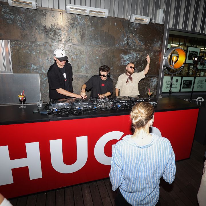 Un DJ si esibisce a un evento all'aperto dietro un bancone rosso con la scritta "HUGO". Tre persone sono al banco del DJ, gli spettatori sono riuniti. Lo sfondo è decorato con elementi industriali e illuminazione calda.