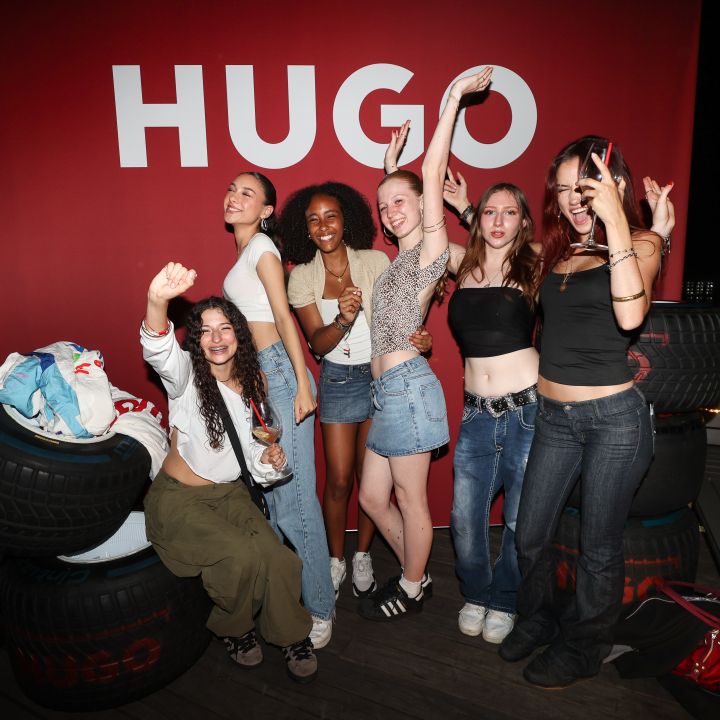 Un gruppo di sei giovani donne posa allegramente davanti a uno sfondo rosso con il testo "HUGO". Sono vestite in modo casual e si trovano tra pneumatici impilati, alcune facendo gesti giocosi con le mani. La scena appare festosa ed energica.