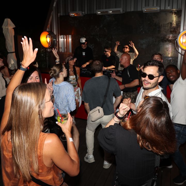 Una vivace scena di festa con persone che ballano e si godono la musica. Diverse persone hanno le mani alzate e sullo sfondo c'è un DJ. Luci al neon e un mix di abbigliamento casual ed elegante contribuiscono all'atmosfera vivace.