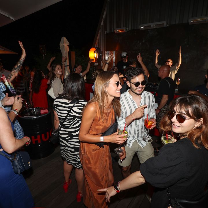 Un vivace gruppo di persone si gode una festa all'aperto di notte. Gli ospiti sorridono, ballano e tengono in mano bevande. L'ambiente è illuminato da una luce calda e ci sono tavoli con botti come supporto. L'atmosfera è festosa e rilassata.