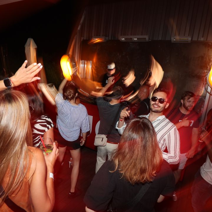 Persone ballano energicamente in un club scarsamente illuminato con illuminazione calda. La sfocatura del movimento cattura l'atmosfera vivace. Una persona in primo piano ha un braccio alzato, mentre altri chiacchierano e si godono la musica.