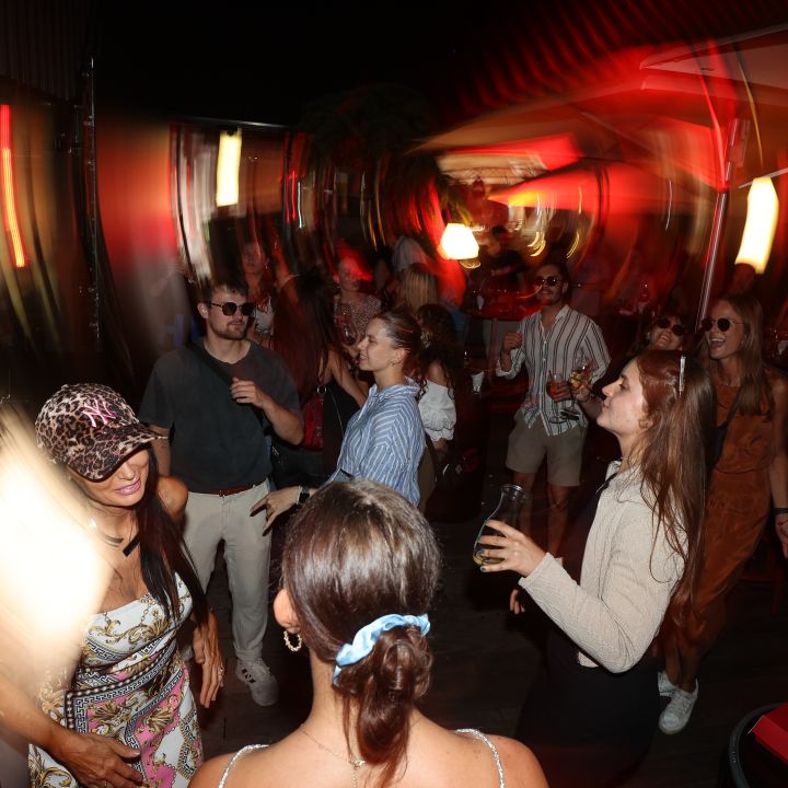 Un'immagine vivida e sfocata di persone che ballano a una festa con luci vorticose e una calda combinazione di colori. Le persone, vestite con abiti estivi casual, sorridono e tengono in mano bevande, creando un'atmosfera di gioia e movimento.