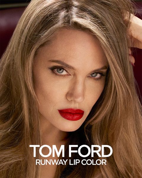 Una persona con i capelli lunghi e un rossetto rosso acceso fissa intensamente. L'immagine è una pubblicità per Tom Ford Runway Lip Color, con il nome del prodotto visualizzato in bianco in basso.