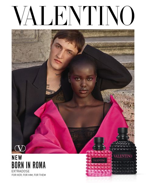 Un uomo e una donna posano insieme su gradini di pietra. L'uomo indossa un cappotto nero, la donna un vistoso abito rosa con spalle scoperte. In basso a destra si vedono due flaconi di profumo Valentino. Sopra c'è scritto "VALENTINO".
