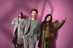 Drei Personen posieren in stilvollen Outfits vor einem rosa Hintergrund. Eine Frau trägt einen karierten Mantel, der Mann in der Mitte einen grauen Anzug, und eine weitere Frau steht rechts in einem grünen Strickpullover und einer Hose.