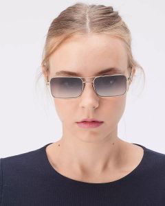 Una persona con capelli castano chiaro indossa occhiali da sole rettangolari e un top blu navy, in piedi davanti a uno sfondo bianco semplice.