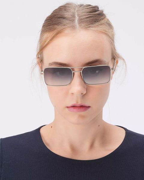 Una persona con capelli castano chiaro indossa occhiali da sole rettangolari e un top blu navy, in piedi davanti a uno sfondo bianco semplice.