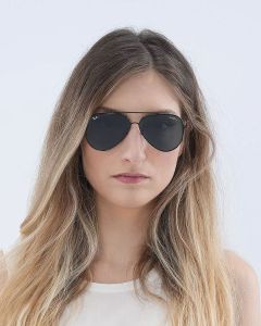 Donna con lunghi capelli biondi ondulati, occhiali da sole da aviatore scuri e top bianco. Ha un'espressione neutra e guarda la fotocamera su uno sfondo chiaro e semplice.