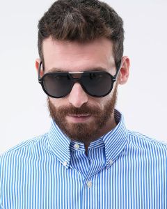 Un uomo barbuto indossa occhiali da sole scuri e una camicia a righe blu e bianche con il colletto abbottonato. Guarda leggermente verso il basso. Lo sfondo è semplicemente bianco.