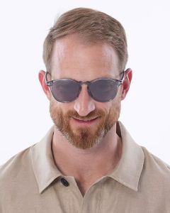 Un uomo con capelli castani corti e barba, occhiali da sole e una camicia beige con colletto sorride davanti a uno sfondo bianco semplice.