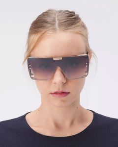 Una persona con i capelli castano chiaro raccolti indossa un paio di occhiali da sole rettangolari oversize con montatura spessa. Indossa un top scuro e semplice e si trova di fronte a uno sfondo bianco semplice.