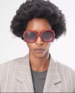 Una persona con un'acconciatura afro di media lunghezza indossa grandi occhiali da sole rossi rotondi e un blazer grigio sopra una camicia scura. Lo sfondo è semplicemente bianco.