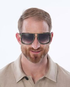 Un uomo con capelli corti e barba indossa un grande paio di occhiali da sole quadrati con lenti scure. Indossa una camicia beige con colletto e sorride. Lo sfondo è semplicemente bianco.