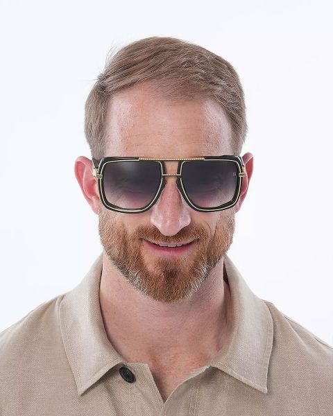 Un uomo con capelli corti e barba indossa un grande paio di occhiali da sole quadrati con lenti scure. Indossa una camicia beige con colletto e sorride. Lo sfondo è semplicemente bianco.
