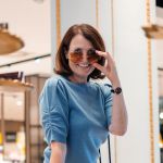Una donna con i capelli castani è all'interno, sorride e solleva un paio di occhiali da sole marroni. Indossa un maglione blu e un orologio e si trova di fronte a uno specchio con elementi di sfondo sfocati.