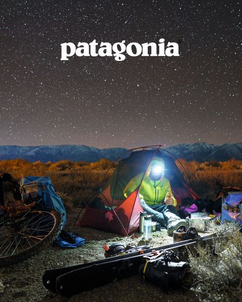 Una persona con una lampada frontale è seduta in una piccola tenda illuminata sotto un cielo stellato vicino a un falò. Nelle vicinanze si vedono biciclette, attrezzatura da esterno e in alto un logo Patagonia. Sullo sfondo si vedono un paesaggio brullo e una catena montuosa.