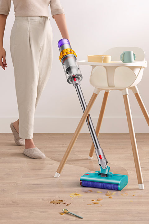 Una persona usa un aspirapolvere senza fili per pulire dei cereali rovesciati su un pavimento di legno accanto a un seggiolone bianco. Una tazza e una ciotola sono sul vassoio del seggiolone. La persona indossa pantaloni chiari e pantofole.