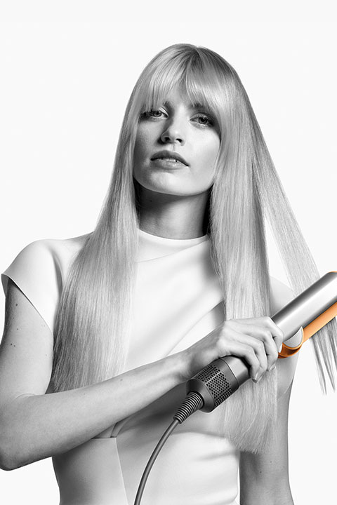 Donna con lunghi capelli biondi che usa una piastra, indossa un top bianco e guarda avanti. Immagine in bianco e nero ad alto contrasto.