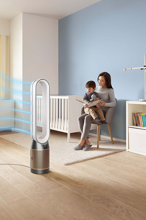 Una donna è seduta su una sedia e legge a un bambino in una stanza dei bambini con culla, libreria e lampada. In primo piano, un purificatore d'aria dal design moderno ed elegante emette flussi d'aria. La stanza ha pavimenti in legno chiaro e pareti blu tenue.