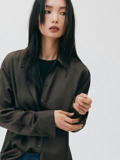 Donna con lunghi capelli neri, che indossa una camicia marrone larga e un top nero, che guarda di lato su uno sfondo bianco semplice.