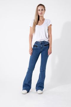 Una persona con una maglietta bianca e jeans a zampa blu è in piedi davanti a uno sfondo bianco semplice. Ha i capelli lunghi e indossa scarpe da ginnastica bianche. Posa con una mano in tasca e l'altra sul fianco.